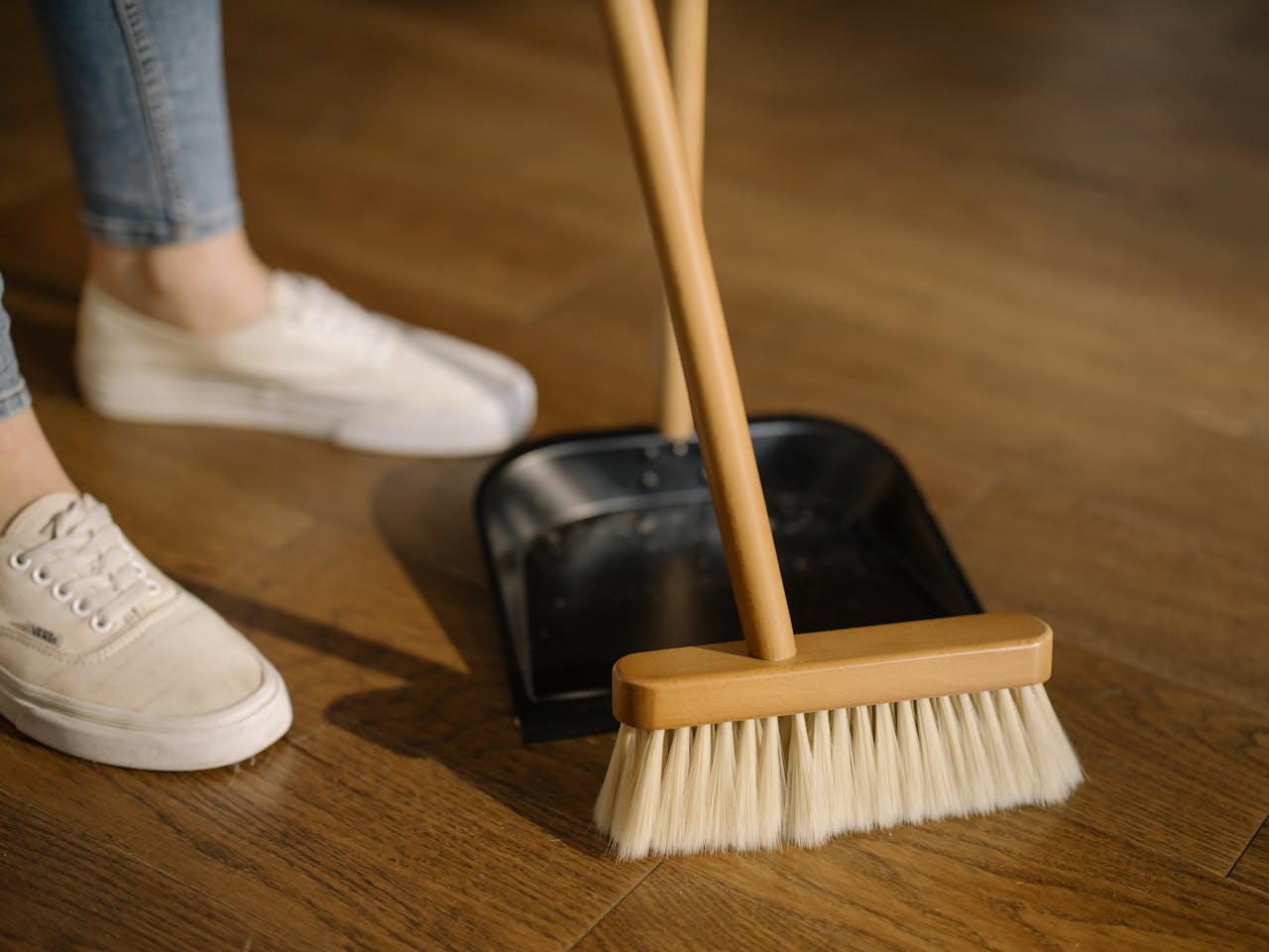 home-hero Close-up of cleaning process with broom and dustpan beside sneakers on a wooden indoor floor.
